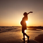 Gezeigt wird eine schwangere Frau als Silhouette an einem leeren Strandabschnitt während des Sonnenuntergangs.