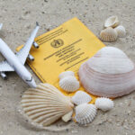 Symbolbild mit einem internationalen Impfausweis (Gelb) Auf feinem Sand mit einigen Muscheln und einem Flugzeugmodell. Gezeigt werden soll die Notwendigkeit den eigenen Impfschutz zu überprüfen beziehungsweise zu ergänzen - vor allem bei anstehenden Fernreisen.