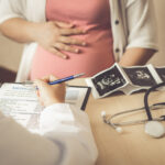 Auf dem Bild erkennbar eine schwangere Frau im Rahmen einer Beratungssituation beim Arzt Vor ihr liegen auf typischem Ultraschallpapier ausgedruckte Untersuchungsbefunde.