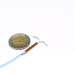 Vergleich der Größe einer T-förmigen Spirale (IUD) aus Kupfer oder Gold zur hormonfreien Verhütung im Vergleich mit einer 2 Euro Münze.