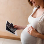 Auf dem Bild wird eine hochschwangere Frau im Querschnitt gezeigt die sich mit der einen Hand den Babybauch hält und in der anderen Hand Ultraschalluntersuchungsbilder betrachtet.