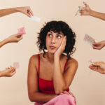 Junge Frau schaut nachdenklich weg, umgeben von Händen, die verschiedene Formen hormoneller und nicht-hormoneller Verhütungsmittel halten. N