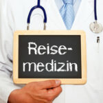 Das Wort Reisemedizin wird auf einer Schiefertafel gezeigt und von einer Person mit Arztkittel und Stethoskop gehalten.