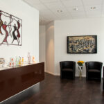 Das Bild zeigt unser Wartezimmer, Mit Fernseher, modernen Stühlen und einem Metallkunstwerk des israelischen Künstlers "JoJo".