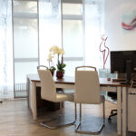 Gezeigt wird unser Sprechzimmer mit 2 Stühlen vor einer großen Fensterfront mit Schalenvorhängen. Ebenso erkennbar ein Skelett und eine weibliche Skulptur des Künstlers "JoJo".