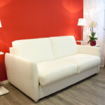 Gezeigt wird eine weiße Ledercouch vor einer Kaminroten Wand mit Stehlampen an beiden Seiten. Rechts zusätzliche Schalenvorhänge und Fenster.