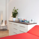 Das Bild zeigt eines unserer Behandlungs und Untersuchungszimmer im Vordergrund eine der Behandlungsligen. Unter angeordnet ein Sideboard mit Schubladen.